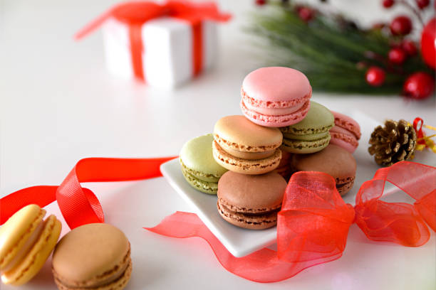 Stack of French macaroons of various flavors on ceramic plate with red bow and gift in the background on white table. Elevated view.