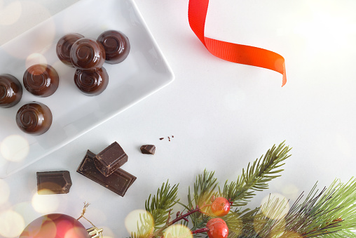White ceramic plate filled with dark chocolate bonbons on a white table with Christmas decorations and portions of choclolate for desserts. Top view.