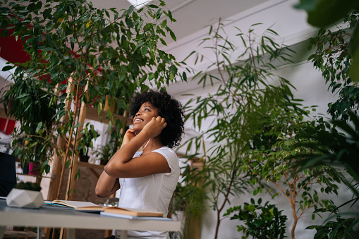 Low-angle view of positive curly African American young woman listening music in earphones sitting at desk with laptop and smiling looking away in light home office room with biophilic interior.