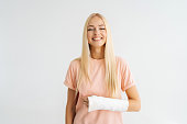 Studio portrait of positive blonde young woman with broken arm wrapped in plaster bandage smiling looking at camera, standing on white isolated background.