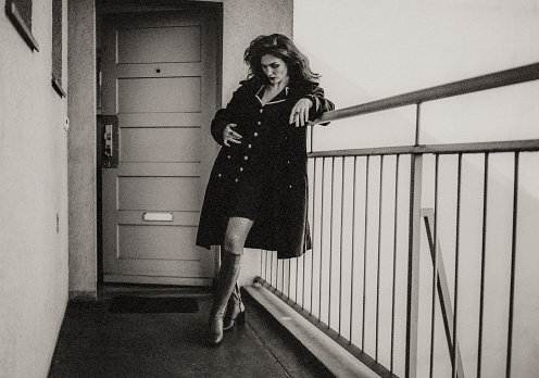 A young, fashionably dressed woman is waiting for someone in front of her open apartment door.