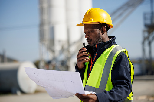 Portrait of industry worker or engineer holding blueprint and using walkie-talkie at industrial facility