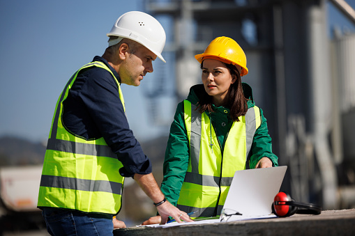 Male and female industrial engineers in hard hats discuss new project while using laptop