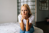 Close-up portrait of pensive young blonde woman sitting alone in living room and sad looking at camera holding hands on chin, thinking over health problems.