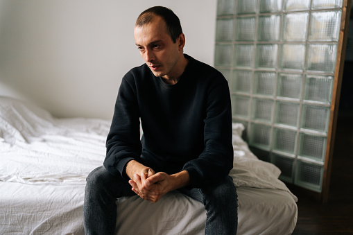 Portrait of desperate male sitting alone at home looking away with pensive facial expression, deep thoughts feeling lonely. Frustrated young man pondering make difficult choice.