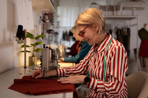 Side view of a happy blonde female fashion designer working with her sewing machine in her shop, putting her creativity to work and making a new project while her female colleague is sitting next to her and helping her