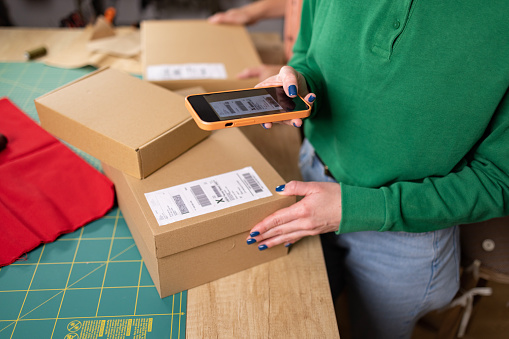 Close up of a smart phone being held by an unrecognizable woman standing next to her workbench taking care of the the packages before shipping them to the clients, using a smart phone to take a photograph of a receipt sticker on the box