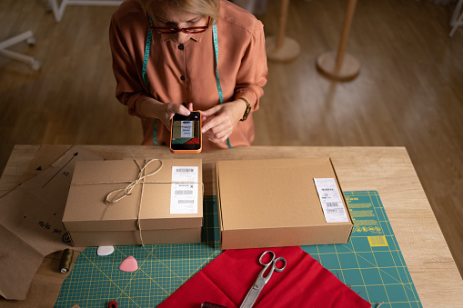 High angle view of a female clothes designer using a smart phone to take scan a bardcode on the packages before sending them to her clients