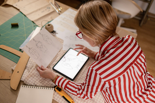 High angle view of a female clothes designer standing next to her workbench full of different tools she uses to create new designs, holding a paper drawing of her sketch and using a digital tablet computer to make corrections