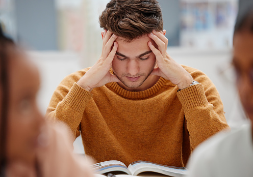 Study, stress and student at university with anxiety for a test. Young man with headache, worried and studying with textbook on desk in library. Focus in education, learning and stressed about exam