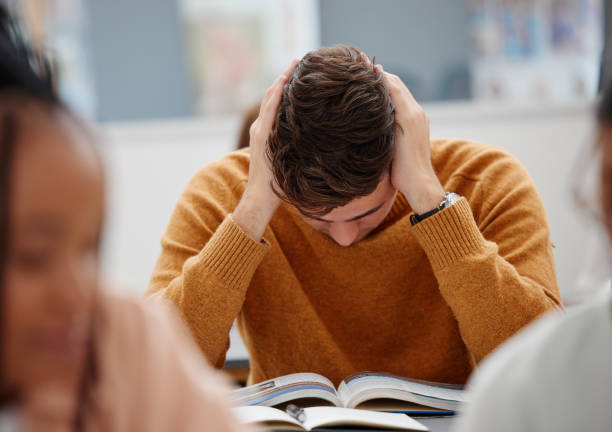 istock University, study and tired student reading book in class while studying for test or exam. Education, stress and burnout man with headache, knowledge and scholarship learning college work in library. 1444197534
