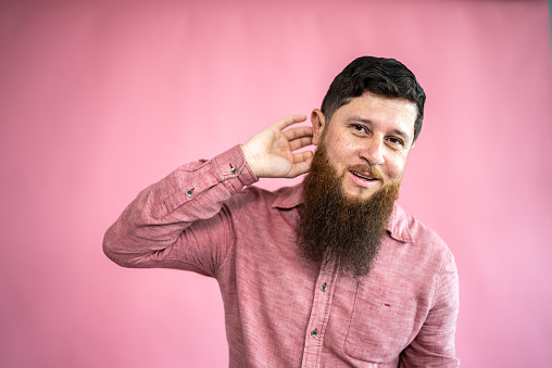 Portrait of a mid adult man putting hand to his ear on a pink background