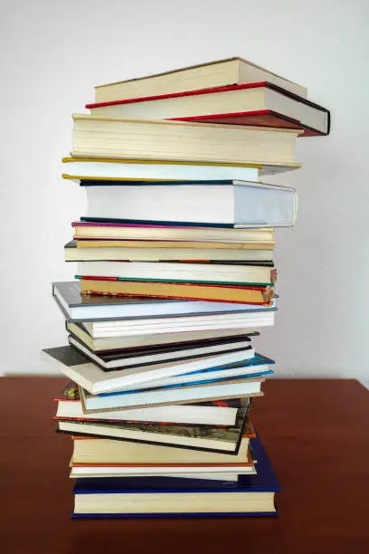 A pile of books of different shapes and thicknesses on a wooden shelf and a white wall in the background