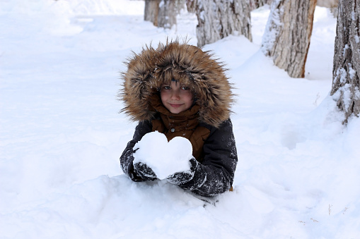 Active games in nature in winter. The boy is buried in a mountain of snow. He holds a heart made of snow in his hands and looks at the camera.