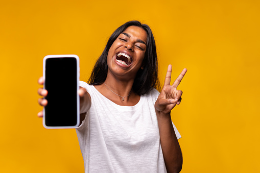 Happy, excited young indian woman showing phone screen to camera doing peace sign gesture with hand. Female looking at camera isolated on yellow background. Technology concept.
