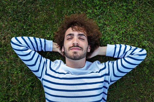 Close up top view of serene, relaxed young man lying down on grass with eyes closed. Wellbeing and lifestyle concept.