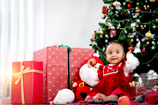 Portrait of adorable happy smiling African American little girl child sitting with many gift boxes presents under Christmas tree in living room, kid celebrating happy Christmas winter holiday