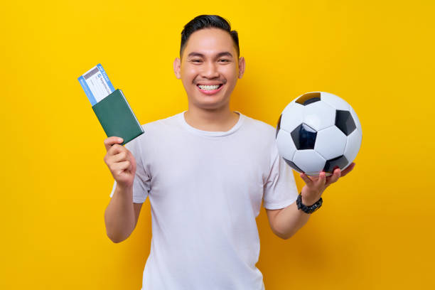 excité jeune fan de football asiatique portant un t-shirt blanc tenant un ballon de football et une carte d’embarquement de passeport isolée sur fond jaune. les gens sport loisir style de vie concept - ticket sport fan american football photos et images de collection