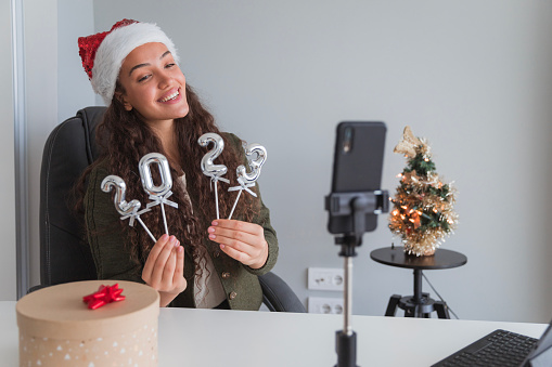 Happy young woman wearing Santa Claus hat vlogging about New Year and holding number shaped balloons