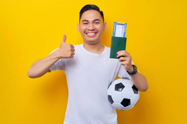 excité jeune fan de football asiatique portant un t-shirt blanc portant un ballon de football et montrant une carte d’embarquement de billet de passeport avec un geste du pouce levé isolé sur fond jaune - ticket sport fan american football photos et images de collection