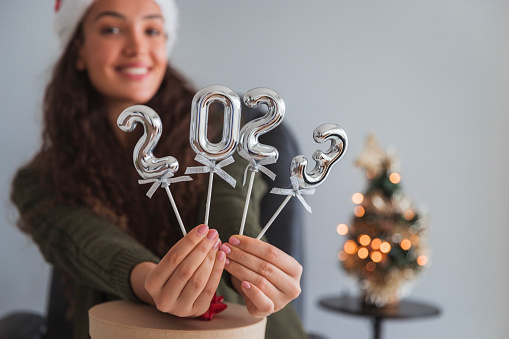 Cheerful young business woman celebrating new year at work, holding balloons shaped as 2023 numbers and wearing a Santa Claus hat