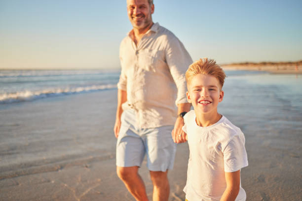 отец и сын сближаются на пляже, гуляют в одиночестве по океану и держатся за руки на закате. портрет взволнованного ребенка, наслаждающегос� - teenager parent beach contemporary стоковые фото и изображения