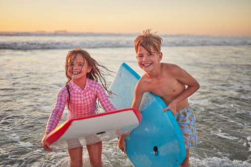 Happy children, beach and learning to surf for fun and bonding on bali summer vacation. Kids, boy and girl siblings on tropical holiday while surfing with board for water sports, splash and swim