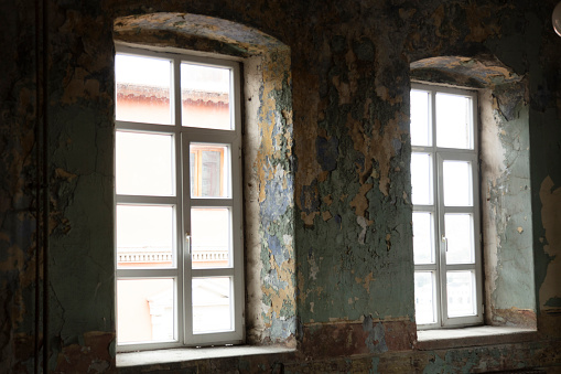 Viewing in an old room with big windows. Empty place in an abandoned building. Bricks come through the wall plaster and paint is peeling off. Ruined apartment in bad condition.