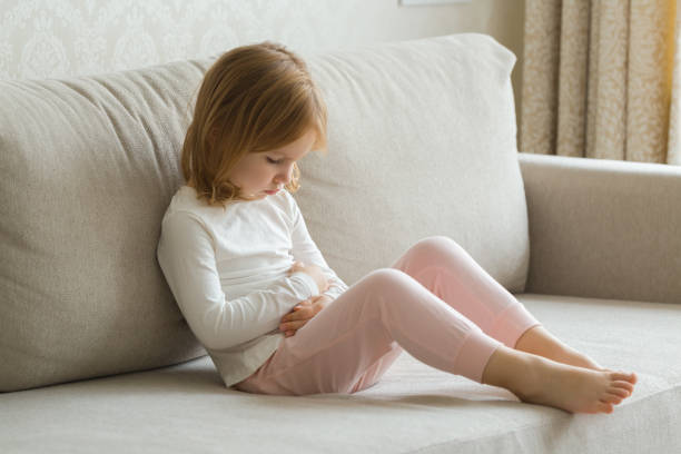 Little girl sitting on sofa and touching painful belly with arms at living room. Child suffering from stomach ache at home. stock photo