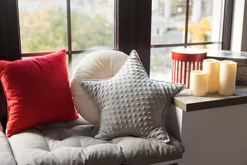 Cozy decorative interior pillows and candles on a wide windowsill by the window for the winter holidays time.
