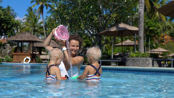 madre e hijos juegan juntos en la piscina con juguetes de natación. mamá sostiene un cubo sobre su cabeza y vierte agua de él, imitando una cascada. consejos para mantener a los niños ocupados durante las vacaciones. - twin tips fotografías e imágenes de stock