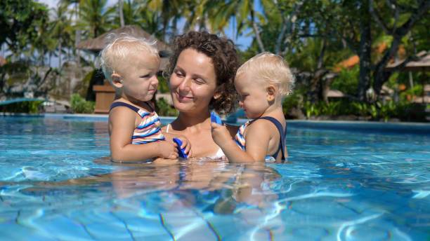 primer plano de una mujer con gemelas, pasan tiempo juntos en una piscina privada. ella los besa uno por uno, mostrando cómo son amados y queridos para ella. - twin tips fotografías e imágenes de stock