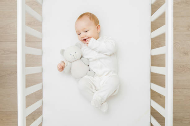 Adorable happy baby boy in white bodysuit hugging teddy bear and lying down on back on mattress in crib at home room. 6 months old infant playing with first friend. Closeup. Top down view. stock photo