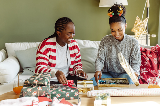 Happy young African-American female friends hanging out at home during the Christmas holidays. They are wrapping Christmas presents and enjoying