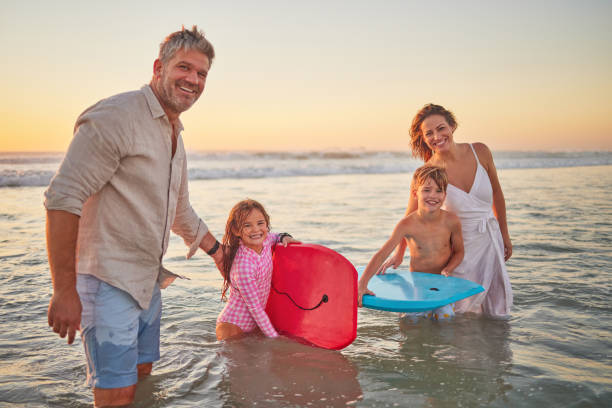 familia, playa y bodyboard, niños con padres en el agua en vacaciones de verano. mamá, papá y niños surfeando en el océano al atardecer en australia. libertad, diversión y vacaciones, hombre y mujer felices juegan en olas - surfing beach family father fotografías e imágenes de stock