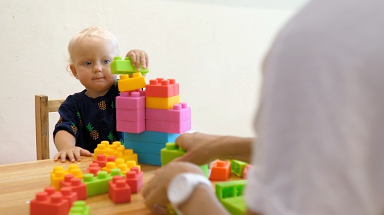 A blond kid builds a tower of colorful plastic building blocks. The kid learns logic and develops fine motor skills of hands together with the teacher. A fun and educational day in kindergarten.