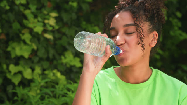 Beautiful mixed race African American girl teenager young woman wearing a green t-shirt outside happy laughing and drinking a bottle of water