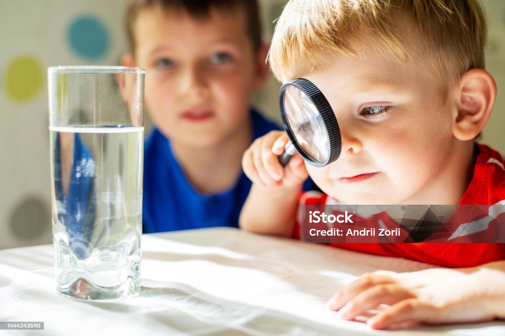 The child boy looking at water in a glass through magnifying glass The child boy looking at water in a glass through magnifying glass. Water quality check concept. STEM - Topic Stock Photo