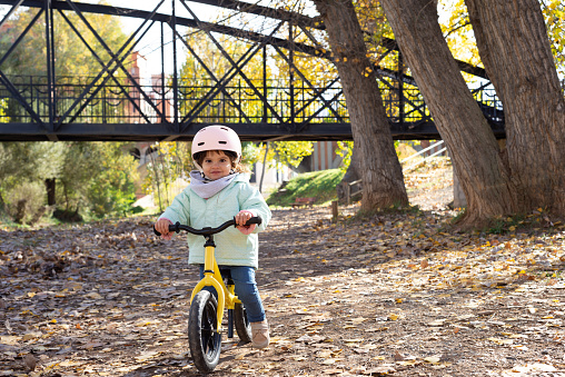 Smiling female toddler wearing a pink cycling helmet riding a balance bike and looking at camera in a park at a sunny day.