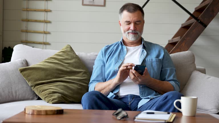 Smiling senior man sitting on couch and using mobile phone at home, chatting