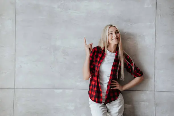 Photo of Attractive Italian blonde woman in plaid shirt pointing aside at empty space against marble wall, satisfied by her health. Fitness concept. Cheerful model at studio. Pretty girl at home. Mockup, sale.