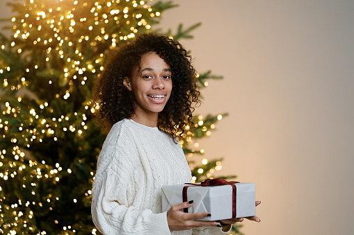 Merry Christmas and Happy Holidays! Young african american woman holding a holiday gift on by the Christmas tree in the cozy living room of the house, the concept of happiness