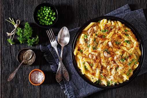 Tuna Mornay, Creamy Tuna Casserole Penne Pasta Bake in baking dish on dark wooden table with fork and spoon, horizontal view from above, flat lay