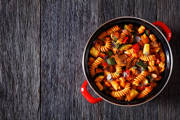 One Pot Vegetable Pasta of rotini, zucchini, capsicum, mushrooms smothered in a garlic herb tomato sauce in red pot on dark wood table , flat lay, copy space, close-up