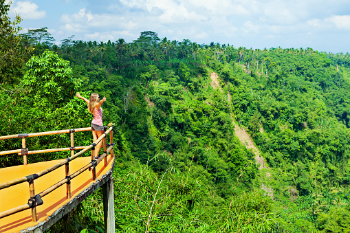 Family vacation lifestyle. Young woman stand on edge of overhanging balcony on high cliff. Happy girl looking at stunning tropical jungle view. Tukad Melangit is popular travel destination in Bali