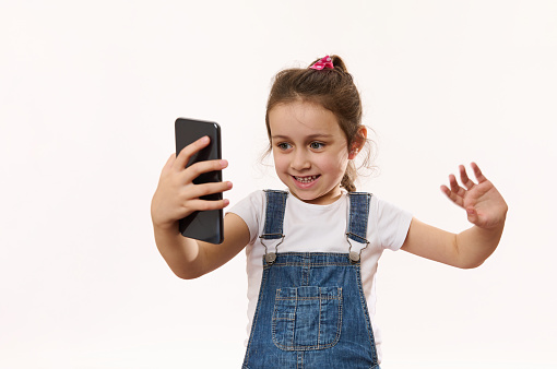 Happy beautiful little girl wearing blue denim overalls, using smartphone, smiling and gesturing hi hello to mobile phone, child communicating with parents on video call, isolated on white background