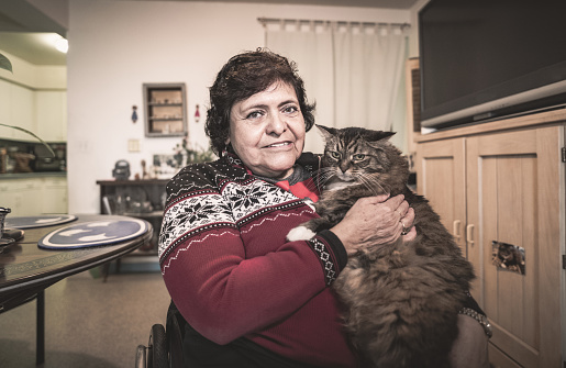 Portrait of the senior disabled Hispanic woman. She is in a wheel chair holding her pet cat, wearing casual clothes. Interior of private apartment.