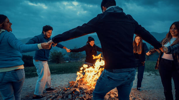 Group of friends having fun together at night party around bonfire - Friendship life style concept with happy people traveler making themselves warm by campfire at dusk night time stock photo