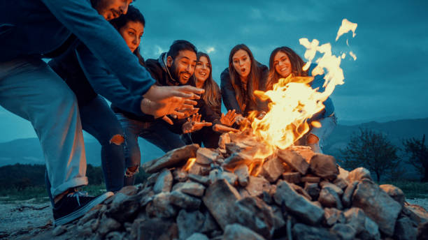 Group of friends having fun together at night party around bonfire - Friendship life style concept with happy people traveler making themselves warm by campfire at dusk night time stock photo