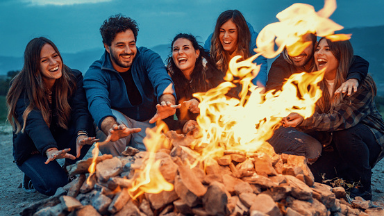 Group of friends having fun together at night party around bonfire - Friendship life style concept with happy people traveler making themselves warm by campfire at dusk night time